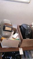 Two welding helmets placed side by side on a table, one black and one silver/gray, with boxes containing drill bits and tools.