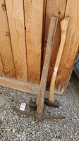 Photo of three yard tools leaning against a wooden fence, showing pickaxe, axe, and sledgehammer with wooden handles and rusty metal heads.