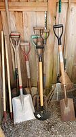 Photo showing the full group of yard tools including shovels, pitchfork, rakes, and spades leaning against a wooden fence.