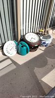 Photo showing outdoor clock, thermometer, turquoise watering can, two ceramic planters on wood trivets, and a resin planter.
