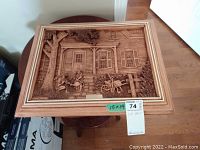 Overall image showing framed bas relief art with rustic house scene, tree, children, wheelbarrow, and some flowers.
