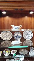 Full display featuring Wedgwood Blue Heritage plates, Johnson Bros Nordic gravy boat with underplate, Delft plate, and Dutch shoe figurines inside a lit cabinet.