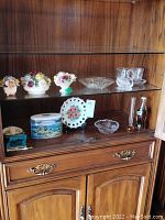 Multiple ceramic floral arrangements, glassware, cream and sugar set, nut dish, swan figurine, and other pieces displayed in a wood and glass cabinet.