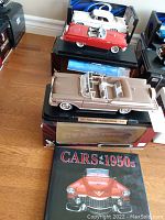 Photo showing three die-cast cars stacked, including the beige 1957 Mercury Turnpike Cruiser, a red convertible, and a white coupe, with the 'Cars of the 1950s' book in front.