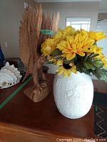 Front view of carved wooden eagle standing on base next to white Kaiser Caprice 317 vase filled with yellow faux flowers including sunflowers placed on table