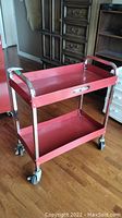 Full view of the red steel rolling shop cart with two shelves and chrome handles on a wood floor.