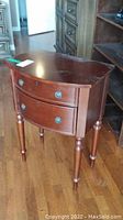 Front left angled view of the curved front two drawer mahogany finish side table with turned legs showing drawers with metal round pulls.