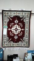 Decorative wall hanging carpet with floral and geometric design in burgundy and cream colors with white fringes, hanging on a rod.