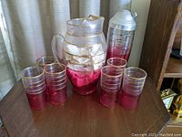 Photo showing large glass pitcher, six matching glasses, and cocktail shaker on wooden surface
