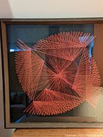 Frontal view of 1970s mid-century modern string art with red strings forming geometric patterns against a black background in a wooden frame.