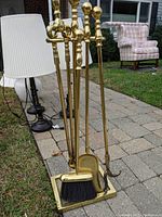 Complete brass fireplace tool set arranged on a sidewalk showing a poker, tongs, shovel, brush on a matching stand.