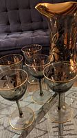 Pitcher with six matched liqueur glasses displayed on a glass table with a gray couch in the background.