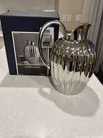 Photo of polished silver Georg Jensen Bernadotte pitcher next to its original branded box on white surface.