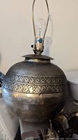 Photo showing a round hammered metal lamp base with decorative geometric patterns on upper half. Includes light socket and harp frame, no lampshade attached.