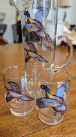 Front view of clear tall glass pitcher and two small glasses with colorful pheasant birds in flight on the front.