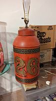 Red metal can lamp with gold Greek Key and number 26 decals, lamp socket fitted with bulb but no shade, some wear visible