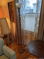 Full view of the tall oak torchiere floor lamp standing next to a window and furniture, showing the entire lamp including the base, spiral pedestal, and top shade.