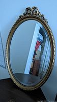 Vertical oval mirror with decorative bow and beaded frame standing upright on a table, reflecting part of a room interior.