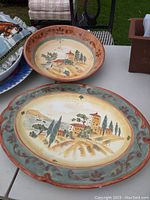 Two large pottery serving platters with Italian countryside landscape scenes, one deeper with handles and brown floral border, other shallower with green-blue border and brown fleur-de-lis pattern.