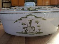 Side view showing green design of a building and trees on white enamel Dutch oven.