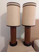 Front view of both lamps with beige fabric lampshades trimmed in brown, placed side by side on a light wood surface.