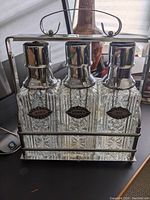 Set of three ribbed glass carafes with metal tops and name tags labeled Scotch, Bourbon, and Rye in metal carrier.