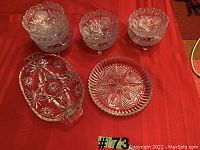 Seven clear glass fruit nappies with scalloped edges and three feet each, plus two decorative glass pickle trays with sections, arranged on red cloth.