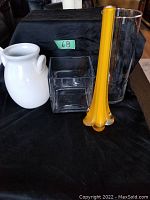 Lot setup showing all four vases arranged on black cloth: yellow bud vase, two clear glass vases (tall cylinder and short square), and white ceramic vase with handles.