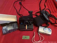 Photo showing Polaroid OneStep SE camera, Olympus digital camera with case, and Arrow-View slide viewer with box all displayed on red fabric background.