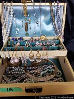 Full view of jewelry box open with numerous necklaces hanging over the top, assorted earrings in slots, rings, pins, and several strands of pearls and chains inside the bottom tray.
