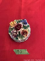 Top view showing delicate multicolored ceramic flower roses on lid with green leaves. Lid stacked on a red fabric surface with wear visible on petals.