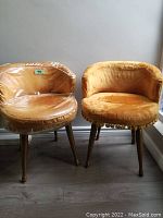 Two vintage barrel back chairs side by side, one with clear vinyl protective cover over plush fabric, the other in exposed plush fabric with tassel fringe.