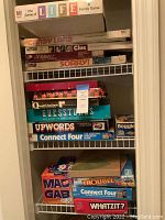Stack of vintage board game boxes on shelving showing titles The Game of Life, Candy Land, Clue, Scrabble, Sorry, Chess, Quad-Ominos, Guesstures, Upwords, Connect Four, Boggle, Mad Gab, Trouble, and Whatzit