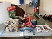 Box with white PVC pipe fittings, electrical cords, caulking guns, painter's tape, toolbox, and toilet repair kits arranged on a table.