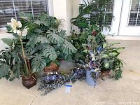 Group of multiple silk plants and flowers arranged on patio floor near window, showing overall collection and containers.