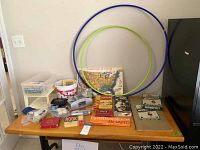 Photo showing collection of vintage board games, puzzles, educational flashcards, card shuffler, and two hula hoops arranged on a table against a wall and beside a refrigerator.