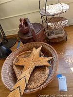 Photo showing large oval wicker basket containing decorative wooden cross and smaller baskets. Round lidded wicker basket with rooster handle, and 3-tier rattan organizer on wooden floor.