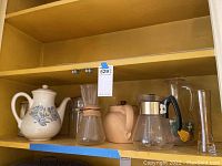 Shelf holding ceramic coffee carafe with floral design, Chemex coffee maker, light brown ceramic pitcher, star-patterned glass carafe with black handle, floral painted glass pitcher, and tall clear glass vase.