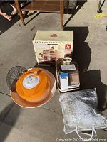 Porcelain enamel cookware box, orange plastic strainer, metal mesh strainer, electric juicer, and cloth bag on a concrete floor.