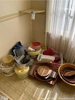 Wide shot of the kitchen items, showing plastic storage containers, plastic and metal bowls, and wood trays stacked in corner of room near window