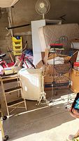 Wide view showing wicker laundry hamper, step stool, multiple plant stands, and other surrounding items in storage setting.