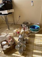 Wide angle shot of the lot showing stacked plates, various mugs, nested bowls, and a teapot. Items are on the floor near a wall outlet.