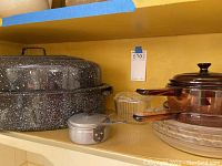 Shelf with two large speckled roasting pans stacked with lids, small metal saucepan in front, and to the right clear glass skillet and Pyrex bowls.