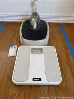 Photo showing both scales together on a wooden floor. One is white with Metro branding and dial. The other is older, metallic, and black with wear.