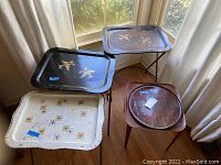 Four vintage trays: Two metal TV trays on foldable legs, one black with floral design and rust, one white with gold/brown design; a small wooden table tray and a round wooden serving tray on floor by window