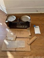 Vintage balance scale inside a worn cardboard box with instruction papers around it, showing signs of use and oxidation on metal scale pans.