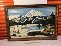 Framed painting of snowy mountain landscape with pond, pine trees, under partly cloudy sky, sitting against orange textured background with ruler for size reference.