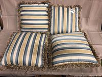 Four decorative pillows arranged on a light brown couch, showing striped pattern in blue, gold, and cream with fringe trim