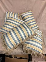 Five decorative pillows with striped satin fabric in blue, gold, and cream, fringed trim around edges, shown stacked on a beige background.