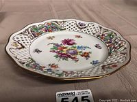 Porcelain dish shown from angled top view on fabric surface, showing the reticulated rim with lattice cutouts and colorful floral patterns along rim and center.